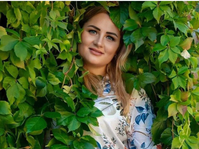 HoneydewWaves, com cabelo loiro, olhos azuis e um sorriso adorável, parcialmente escondida por trepadeiras verdes, vestindo uma camisa com estampa floral, com uma aparência relaxada e bonita..