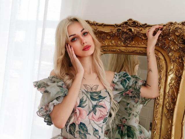 LaraMoss with blonde hair, floral dress, hands on cheeks, gold mirror, butterfly tattoo on chest, pink lips, tattoos on arms, standing near white curtain, looking at mirror.