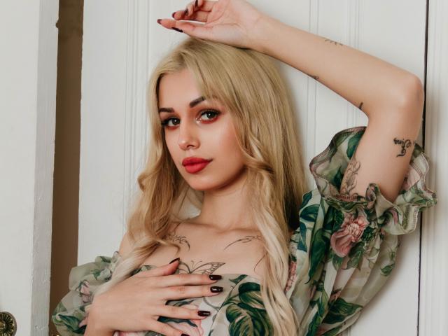 Blonde LaraMoss with red lips, floral dress, tattoos on chest and arm, hand on tits, arm raised, nails painted dark, looking at camera, soft light, slightly parted lips.