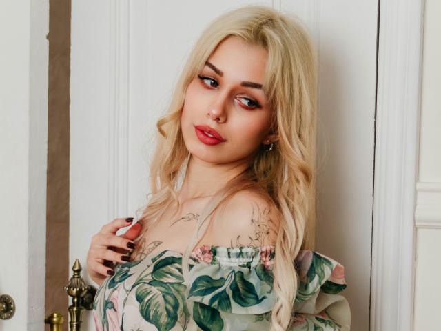 LaraMoss with blonde hair, red lipstick, and floral dress, looking down, hand on chest, black nails, nose ring, tattoo on chest, soft makeup, white door background.