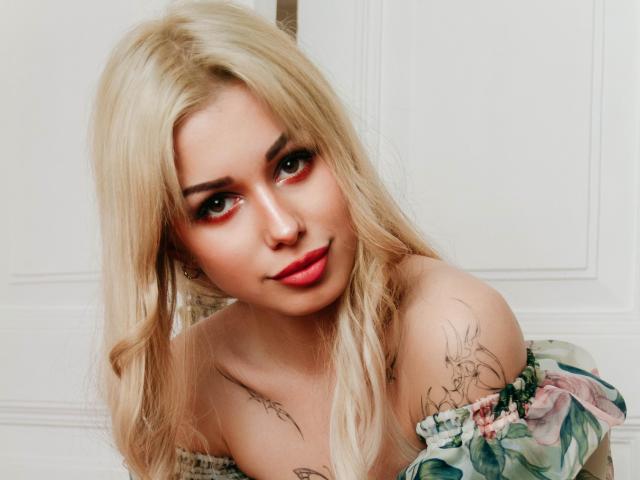 LaraMoss with blonde hair, red lipstick, and floral off-shoulder top, showing tattoos on her shoulder and arm, giving a slightly smirking, seductive look.