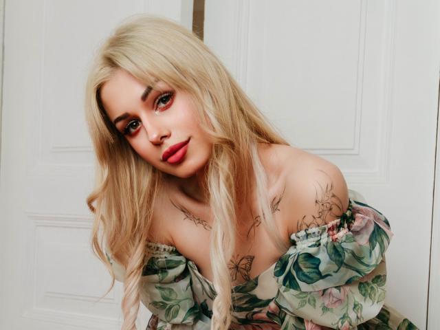 LaraMoss with long blonde hair, wearing a floral off-shoulder dress, has tattoos on her chest and upper arms, and is leaning forward with a cute, slightly seductive look.