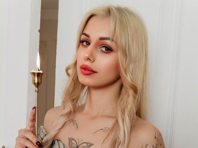 Blonde LaraMoss with wavy hair, red lipstick, tattoos on chest, holding candle, looking at camera, showing off small tits. White wall in background. Soft light.