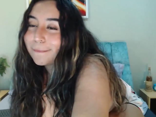 SalomettX has long, wavy dark hair with blonde highlights, a nose ring, and a slight smile, sitting on a bed with a blue headboard in the background.
