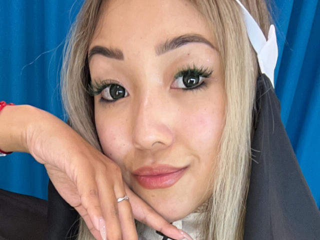 RenyLime with blonde hair, green eyes, and long lashes, wearing a nun outfit, smiling with her hand near her face, showing a ring on her finger.