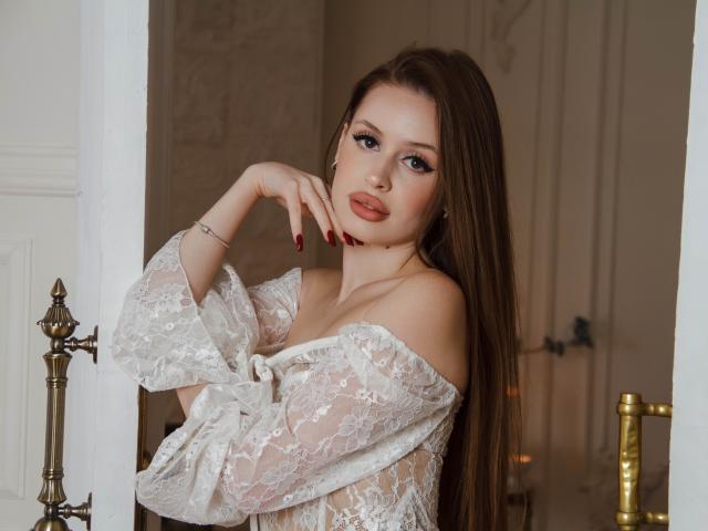 CherylFoxy with long brown hair, wearing a white lace off-shoulder top, posing with one hand on her chin, red nails, and a seductive look, in a doorframe.