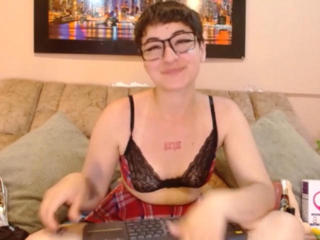 MissTat with short black hair and glasses, wearing a black lace bra and red plaid skirt, typing on a laptop, smiling with a tattoo on her chest.