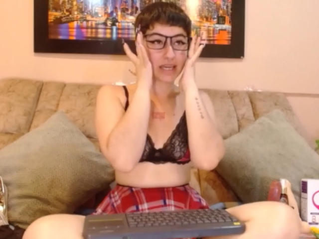 MissTat with short dark hair and glasses, wearing a black lace bra and red plaid skirt, sitting on a couch with hands on her cheeks, laptop in front.