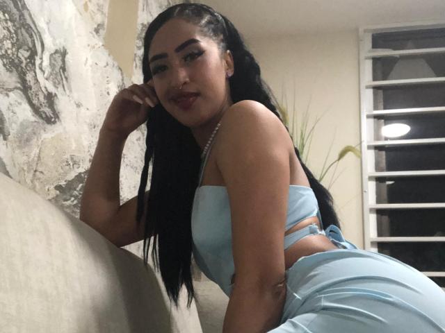 MichellDiaz, con largas trenzas negras, vestida con un top corto azul claro y pantalones a juego, apoyada en un sofá, mostrando sus pechos y una linda sonrisa, en una habitación con poca luz..