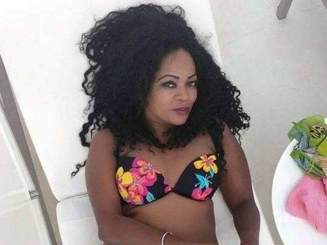 FabioSexy with dark curly hair, brown skin, and large tits in a black floral bikini, lying on a white chair, looking at the camera with a slight smile.