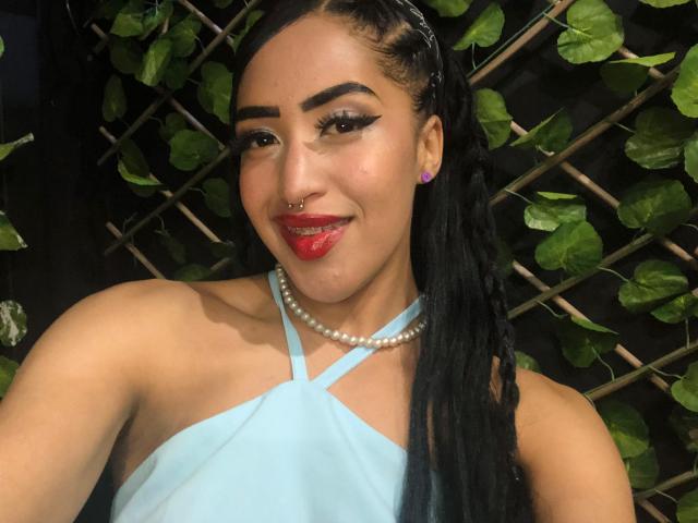 MichellDiaz hat lange, schwarze Zöpfe, roten Lippenstift, einen Nasenring, eine Perlenkette und ein hellblaues Kleid mit gekreuzten Trägern. Sie lächelt mit herausgesteckter Zunge. Sie ist süß und sexy..