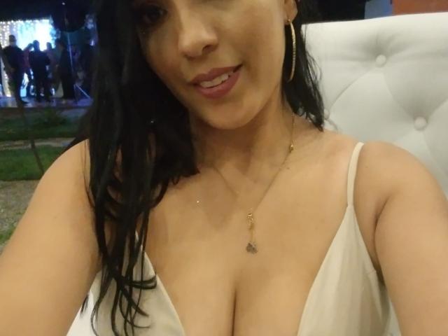 ThamaraStar with large, sexy tits in a low-cut white top, black wavy hair, gold hoop earrings, and a delicate necklace, smiling at the camera.