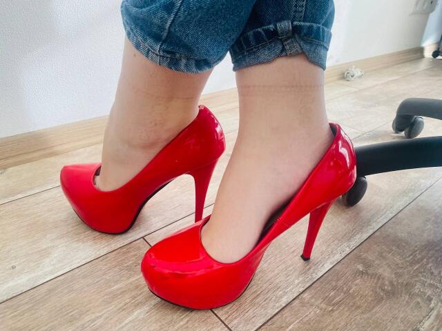 Steisii's shiny red high heels with pointed toes and stiletto heels, standing on wooden floor, wearing blue jeans with cuffs rolled up. Visible office chair on right.