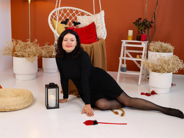 LolaHikls with black turtleneck and skirt, black pantyhose, red headband, sitting on white floor, legs spread, lantern, red rose, white chair, orange wall, curly brown hair, smiling.