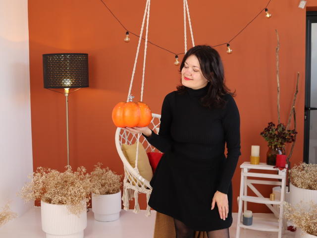 LolaHikls con cabello oscuro y ondulado, vestido negro de cuello alto, sosteniendo una calabaza naranja, de pie en una habitación acogedora, con una silla colgante blanca, una lámpara negra, una mesa blanca con plantas secas..