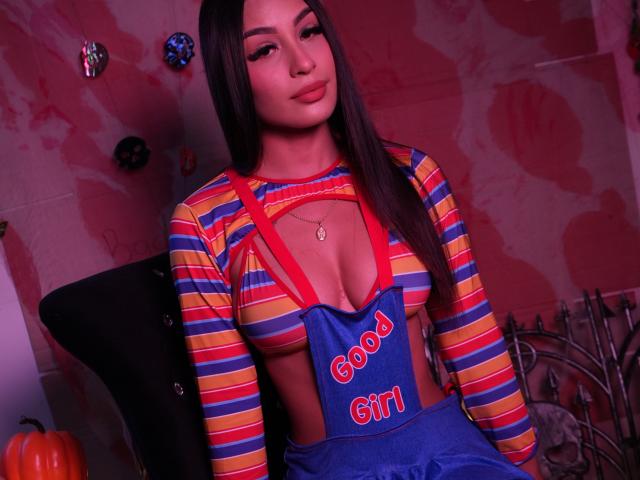 MiaFisher with long dark hair, wearing a colorful striped top, blue suspenders with "Good Girl" text, and a red necklace, showing her cleavage, in a pink-lit room.