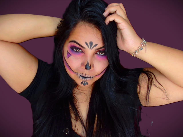 AvaChanne with dark makeup, purple eyeshadow, black nose, and skull face paint, raising arms, wearing black top, purple background, silver bracelet, long black hair.