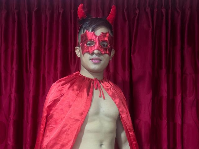 DayanWilly con maschera da diavolo rossa, mantello rosso e corna da diavolo, in piedi senza camicia, che mostra un petto muscoloso e nudo, e rossetto rosa. Sfondo con tenda rossa..