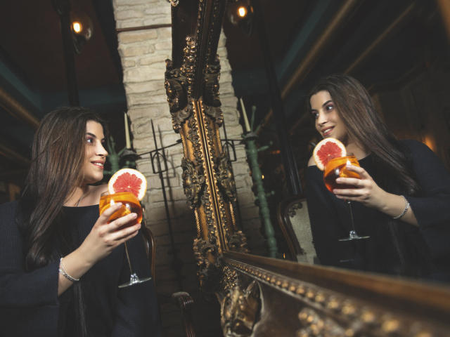 JessieBlaze with long brown hair, black top, holding pink grapefruit cocktail in glass, reflected in ornate gold mirror, smiling at her reflection, brick wall in background.