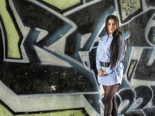 JessieBlaze with long brown hair, wearing a light blue dress, black tights, and a brown belt, standing against graffiti, hands in front, smiling subtly.