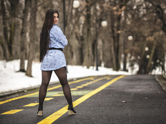 JessieBlaze stands on a snow-lined road, wearing a blue patterned dress, black tights, and wedge shoes, with long dark hair, looking back confidently. Her ass is prominent.