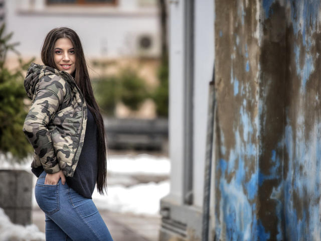 JessieBlaze with long brown hair, smiling, wearing a camouflage jacket, blue jeans, and a black shirt, standing by a weathered blue wall, showing a small butt.