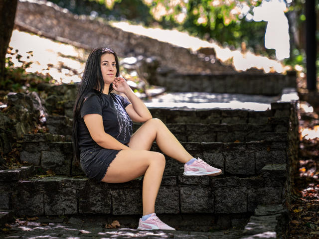 JessieBlaze with long black hair, wearing a dark gray T-shirt and short denim skirt, sitting on stone steps, legs crossed, pink and white sneakers, looking to the side.