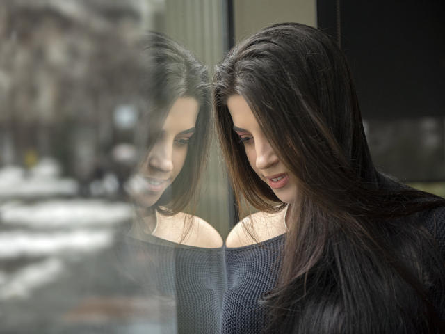 JessieBlaze with long black hair, looking down, wearing an off-shoulder dark blue sweater, reflection in glass, subtle smile, soft natural light, gentle expression, subtle beauty.