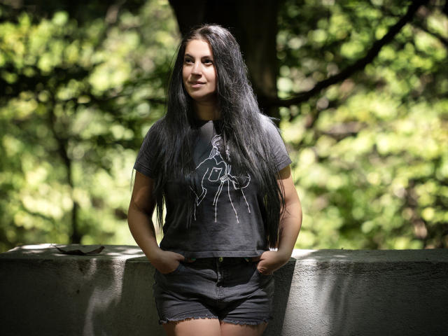 JessieBlaze with long black hair, dark gray t-shirt, and black shorts, hands in pockets, standing against a gray wall, forest background, smiling, natural light, casual style.