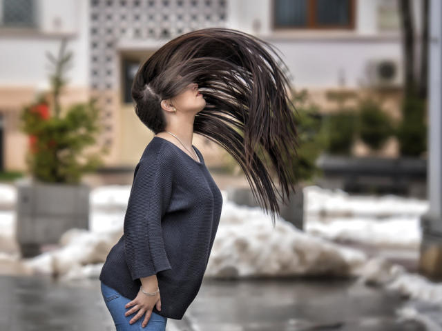 JessieBlaze with long, flying brown hair, wearing a dark blue sweater and blue jeans, standing sideways, hands on hips, in an outdoor urban setting. Her ass is visible.
