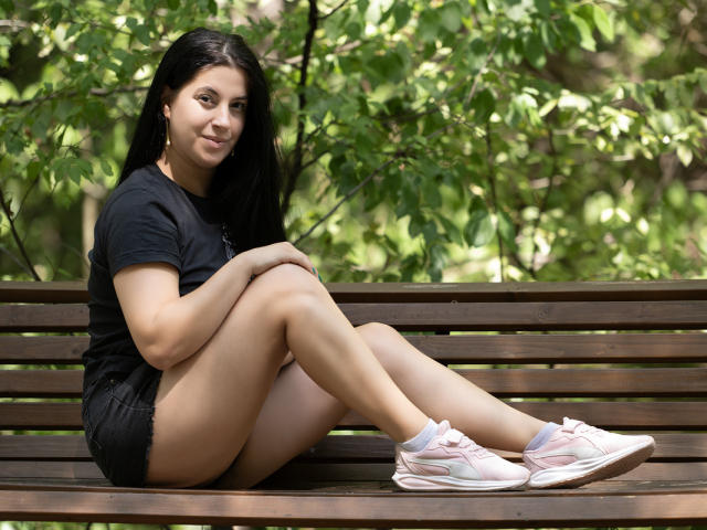JessieBlaze sits on a wooden bench, legs crossed, wearing a black t-shirt, short black shorts, and pink sneakers, with long black hair and a subtle smile.