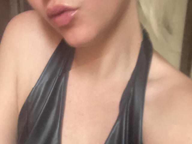BellaBella69s with glossy pink lips, wearing a shiny black halter top, showing her bare shoulders and part of her chest. Her skin is fair, and she's slightly tilted.
