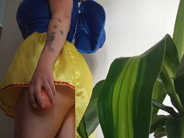 JeneKurz's yellow skirt, blue top, and visible buttcheek with a hand on her pussy. She has a tattoo on her arm and a blue bag. Green plant on the right.