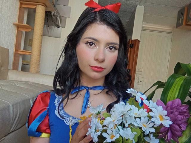 JeneKurz with black hair, red bow, blue and red dress, holding white daisy bouquet, in living room. Cute face, small tits, fair skin, red lipstick.