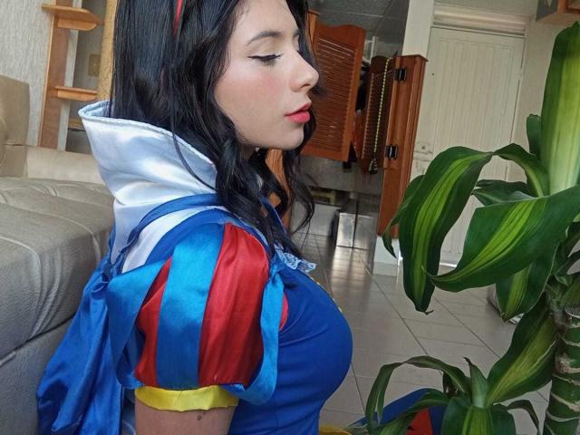 JeneKurz with big tits in a blue and red Snow White cosplay, blue dress, white collar, black wavy hair, red lipstick, standing by a green plant, side profile.