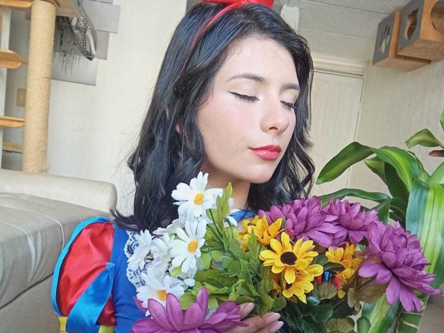 JeneKurz, com cabelo preto ondulado, faixa vermelha na cabeça, vestido azul e vermelho, segurando um buquê de flores roxas e amarelas, flores brancas e amarelas, lábios vermelhos, maquiagem suave, fofa..