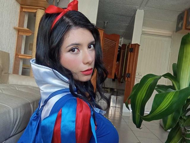 JeneKurz in a blue and red cosplay outfit with a red bow, looking over her shoulder, showing her tits in a bright indoor setting.