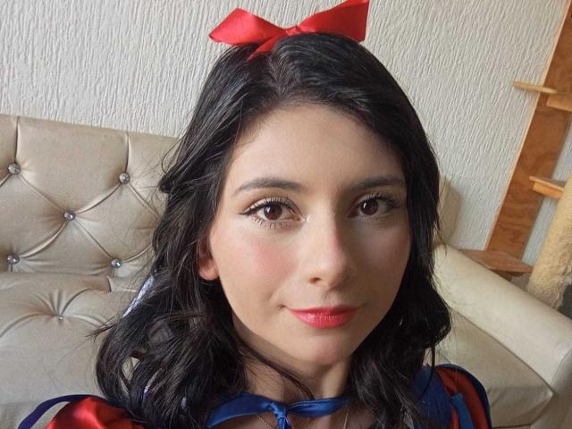 JeneKurz with dark wavy hair, red bow, and eyeliner, wearing a blue and red dress, showing a cute smile, with pink lips, and brown eyes.