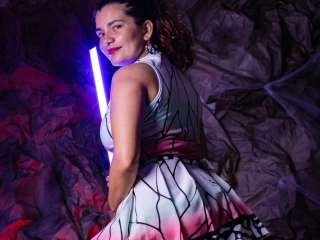 BulmaHot with curly brown hair, wearing a sleeveless, geometric-patterned dress, standing by a purple neon light, looking over her shoulder, showing her ass, smiling with red lipstick.
