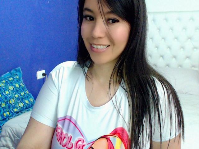 LizBellaa with long black hair, smiling, wearing a white t-shirt with a pink heart and text, sitting on a bed, showing her cute, sexy face.