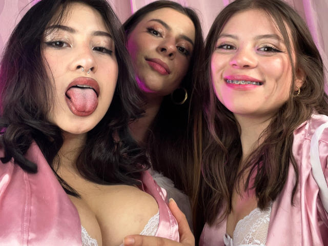Three VictoriaAngels with their tongues out, wearing pink satin robes, showing cleavage in white lace bras. One with a nose ring, all smiling. Pink and purple lighting.