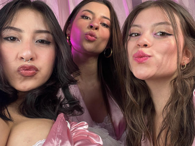 Three VictoriaAngels with pouty lips, dark to light brown hair, and pink lipstick, wearing pink satin and lace, showing their cleavages, with a nose ring and gold earrings.