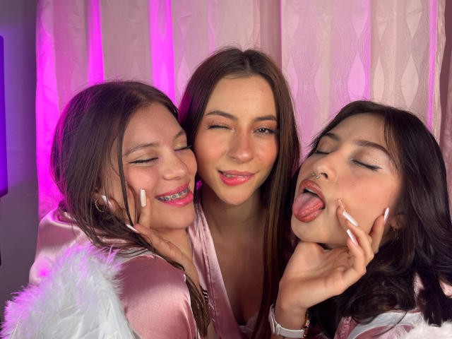 Three VictoriaAngels with pink makeup, one winking, one smiling, and one sticking out tongue, wearing pink satin and white fur, in front of pink-lit curtain.