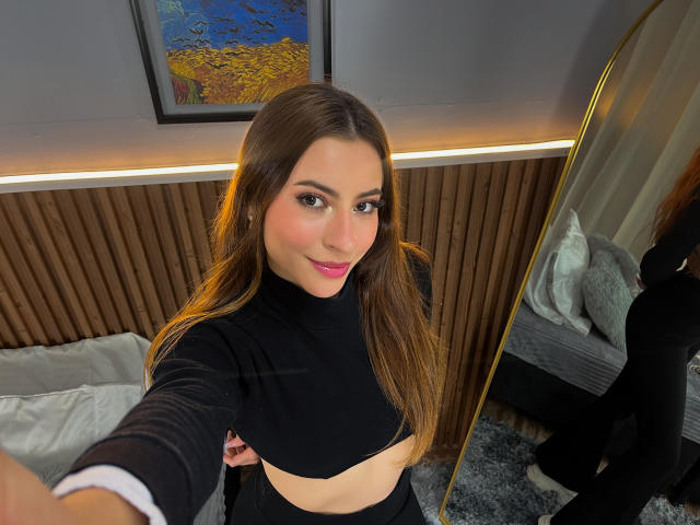 VictoriaAngels with long brown hair, pink lipstick, black crop top, and black pants, smiling at the camera, reflected in a mirror, showing her small tits and slim figure.