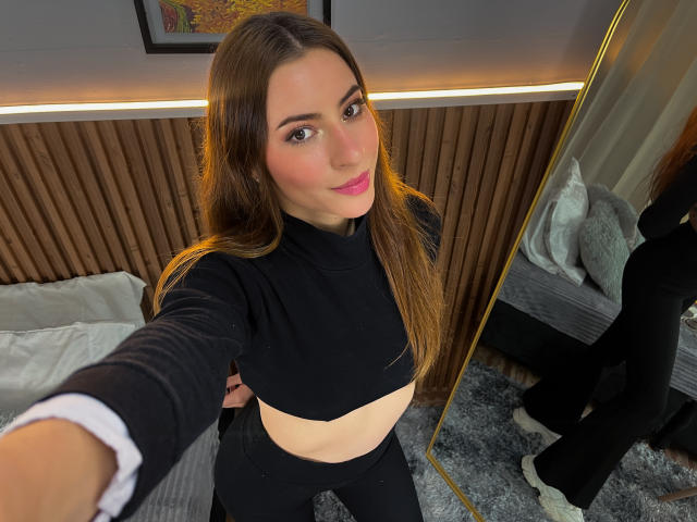 VictoriaAngels has long brown hair, pink lipstick, black crop top, and black pants, taking a selfie with a mirror reflecting her lower body and a bed in the background.