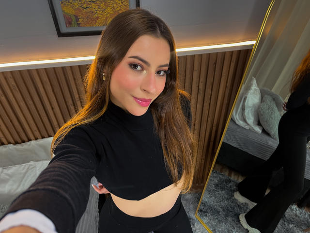 VictoriaAngels with long brown hair, pink lipstick, black top, showing her bare tits, holding phone for selfie, standing in front of mirror, wood-paneled wall, and bed in background.