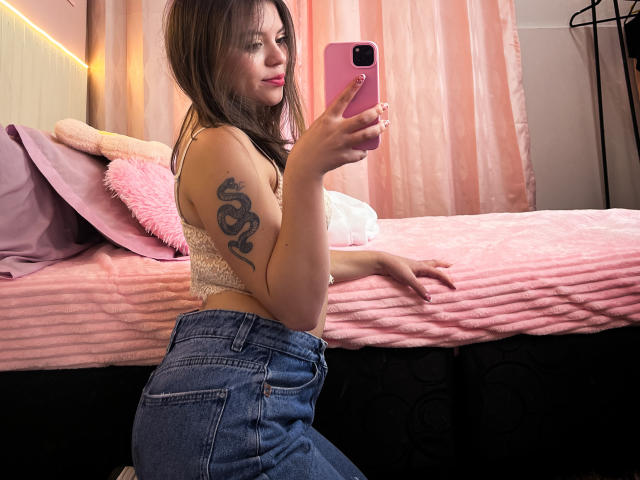 VictoriaAngels with a snake tattoo on her arm, wearing a lace top and high-waisted jeans, kneeling by a pink bed, taking a selfie with a pink phone.