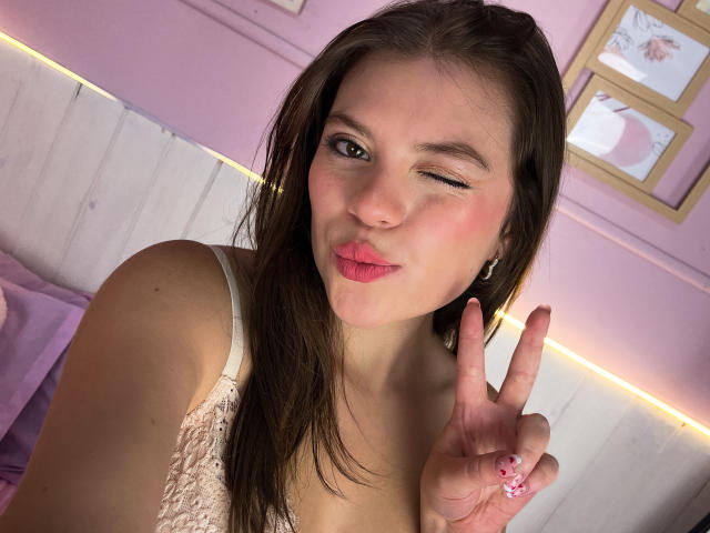 VictoriaAngels with pouty pink lips, winking, making a peace sign with her right hand, wearing a white lace top, brown hair, and pink nails, in a pink room.