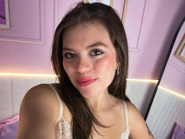 VictoriaAngels has a cute face with pink lips, brown eyes, and long brown hair, wearing a lace top, and a white hair clip. Her small tits are barely visible.