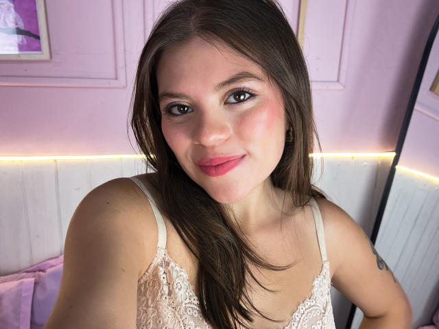 VictoriaAngels has pink lipstick, brown eyes, and long brown hair. She's wearing a white lace top, showing her small tits and a tattoo on her right arm.